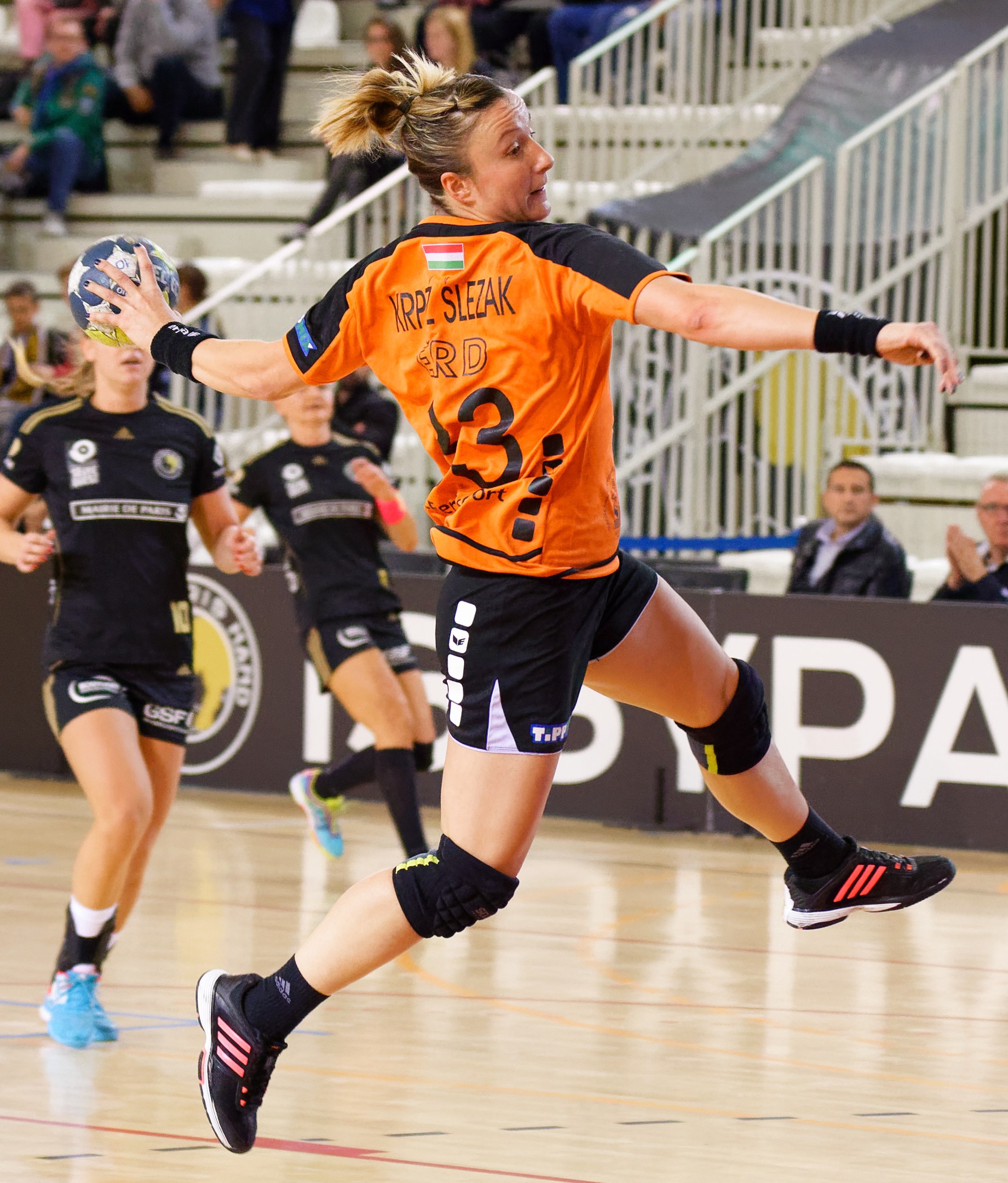 Eine Frau in einem orangen T-Shirt und schwarzen Shorts hält einen Handball, mit Zuschauern und einem "Krp Slezak - Handball-Weltmeisterschaft 2019"-Schild im Hintergrund.