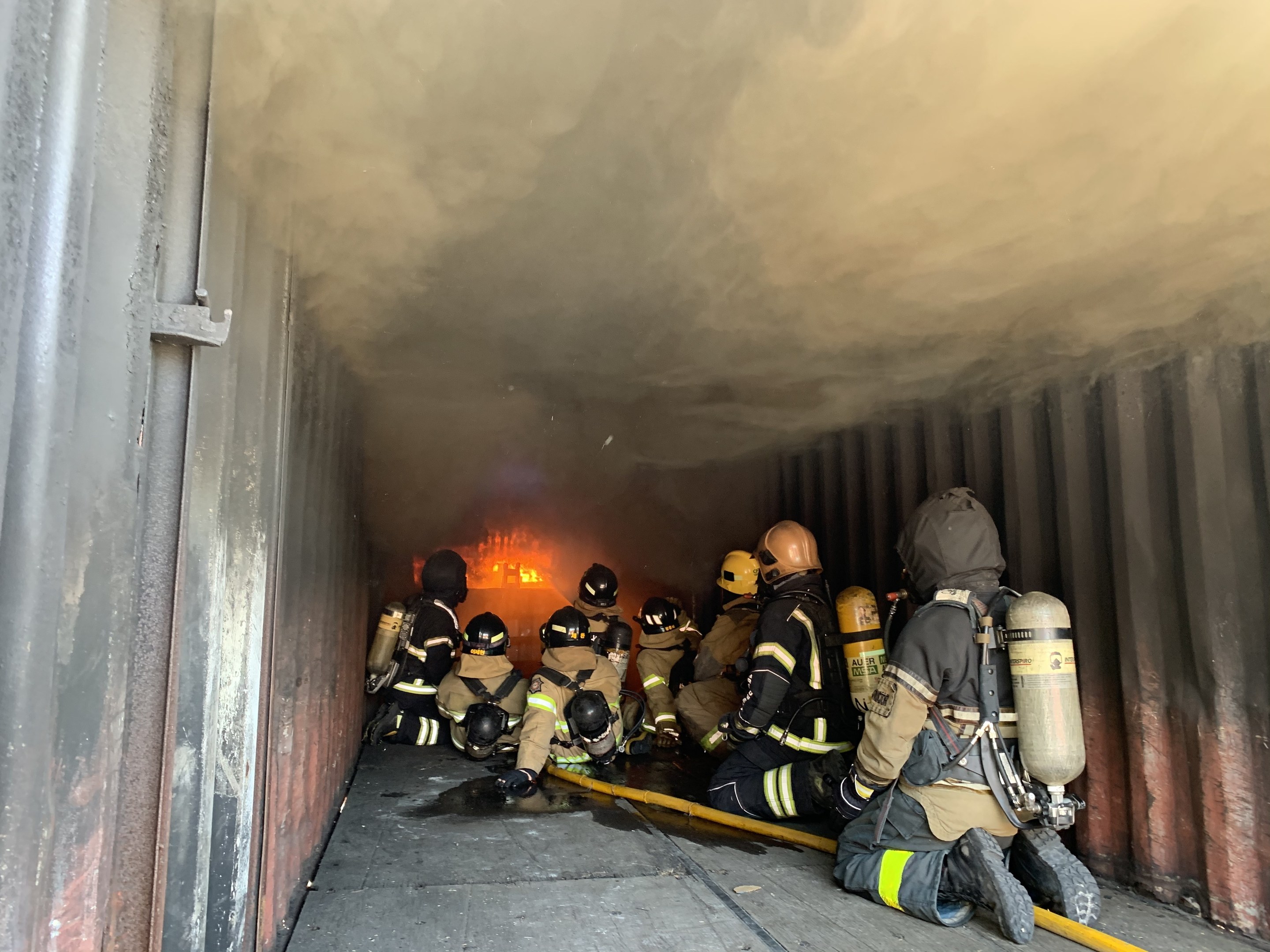 Feuerwehrleute mit Helmen und Sauerstoffgeräten betreten einen raucherföllten Tunnel, einer hölt ein Rohr, mit einem sichtbaren Feuer und einer Wand links.
