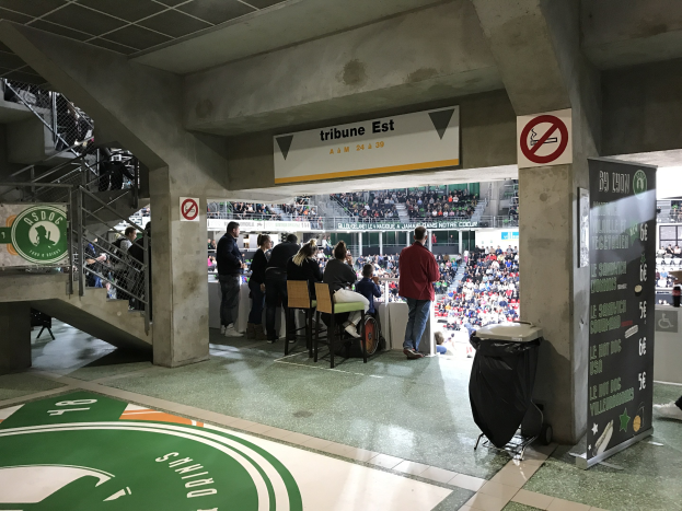 Eine Gruppe von Menschen steht vor einer sitzenden Menge in einem Stadion, mit Schildern, die "Boston Celtics vs New York Jets" anzeigen, einem Müllcontainer, einer Tafel mit Text, einer Treppe mit Geländern und einer Decke mit Deckenleuchten.