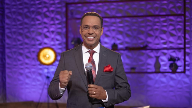 Stephen Colbert, Moderator von *The Daily Show with Trevor Noah*, steht auf einer Bühne und hält ein Mikrofon in der Hand, trägt einen Anzug, eine Krawatte und eine Brille, während im Hintergrund ein Scheinwerfer und ein Ständer zu sehen sind.