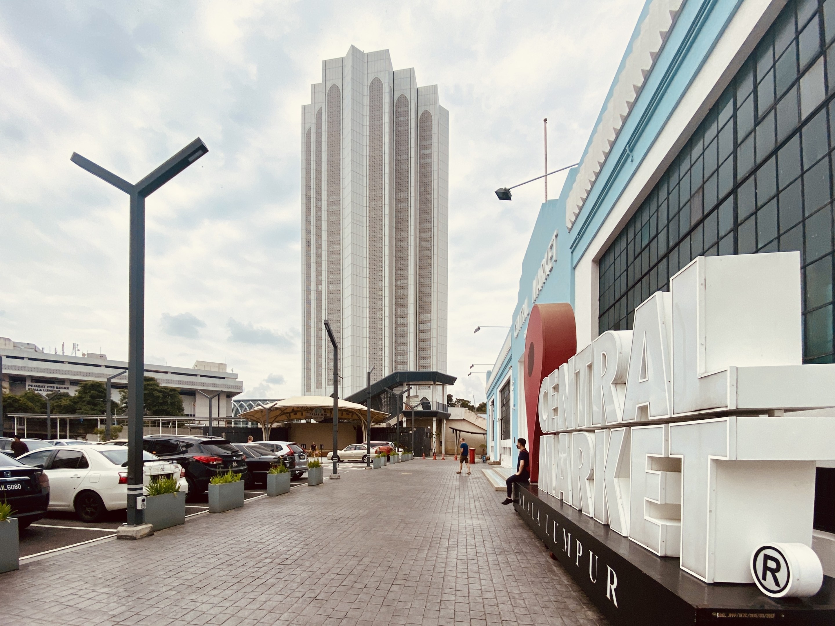 Ein belebter zentraler Markt in Kuala Lumpur, Malaysia, mit hohen Gebäuden, Straßenlaternen, Fahrzeugen, Fußgängern, Schildern, Bäumen und einem bewölkten Himmel.