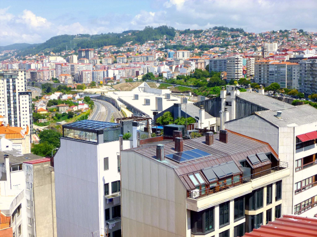Eine Stadtansicht von einem hohen Standpunkt aus, die zahlreiche Gebäude, Bäume, Straßen, Fahrzeuge und ferne Berge unter einem bewölkten Himmel zeigt, wobei auf dem Dach eines Gebäudes Solarpaneele zu sehen sind.