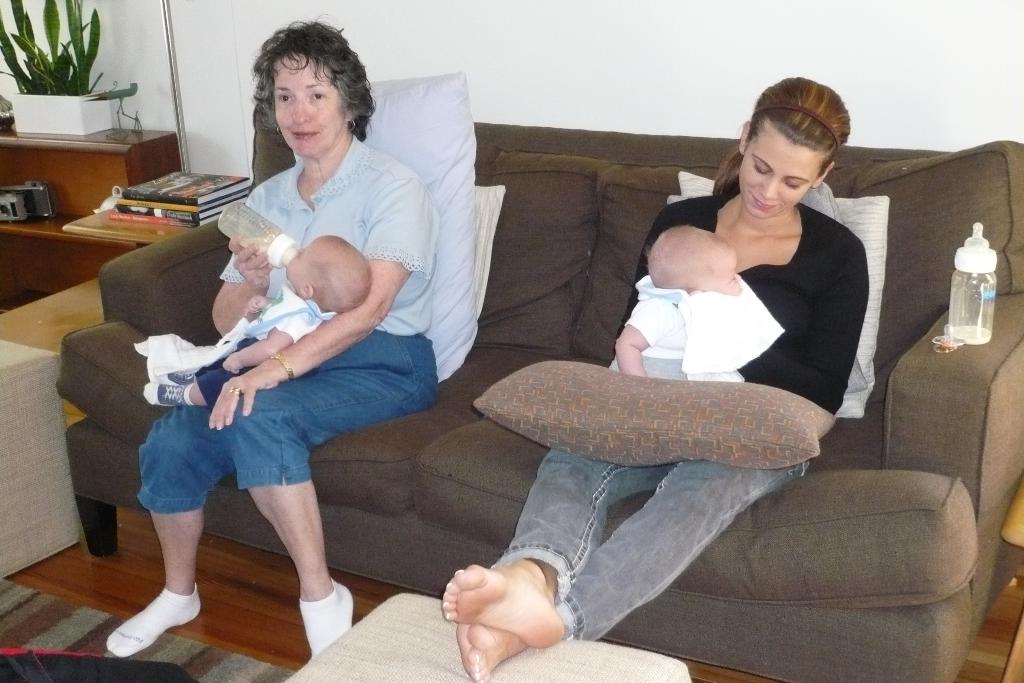 Zwei Frauen sitzen auf einem Sofa und halten Babys, mit Kissen drumherum, vor einer weißen Wand, mit einem Schreibtisch, Büchern und einer Zimmerpflanze links.