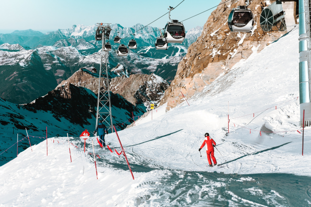 Gruppe von Skifahrern, die eine schneebedeckte Abfahrt mit einem Seilbahnwagen im Hintergrund hinunterfahren, unter einem klaren blauen Himmel mit einer Bergkette.