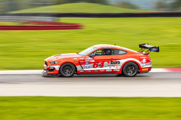 Ein Ford Mustang GT Rennauto mit Text und Zahlen auf der Karosserie fährt auf einer Rennstrecke, mit einem Helmträger im Sitz, Hintergrund unscharf, um Geschwindigkeit und Bewegung anzudeuten.