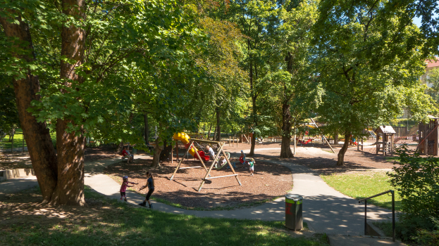 Ein Park mit einem Spielplatz in der Mitte, umgeben von Bäumen, mit Kindern, die auf Geräten spielen, einem Mülleimer, Geländern, Pflanzen, Gras und Gebäuden im Hintergrund unter einem klaren blauen Himmel.