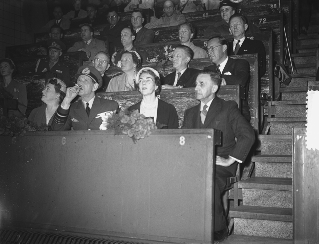 Schwarzes und weißes Foto einer Menge, die auf einer Tribüne sitzt und einer Bühne mit einem Blumenstrauß gegenübersteht, Treppe auf der rechten Seite sichtbar.