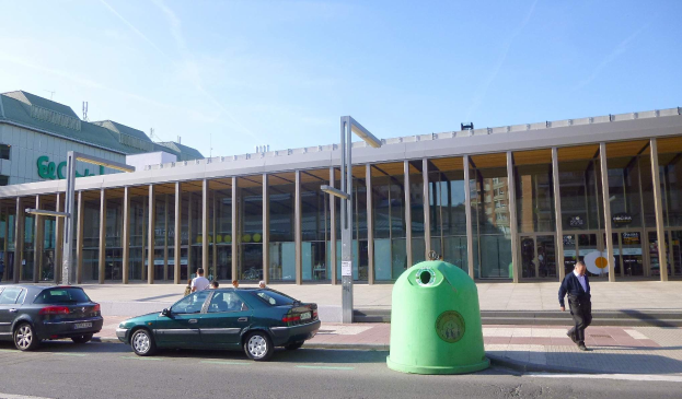 Grüner Müllcontainer an der Seite einer Straße neben einem Glaswandgebäude mit Säulen, mit Fahrzeugen und Fußgängern in der Nähe und einer bewölkten Kulisse am Berliner Hauptbahnhof.