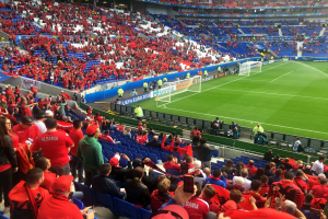 Eine große Menschenmenge schaut einem Fußballspiel in einem Stadion zu, mit Menschen auf Stühlen und Stehplätzen, Fahnen, einem Torpfosten und Deckenleuchten.