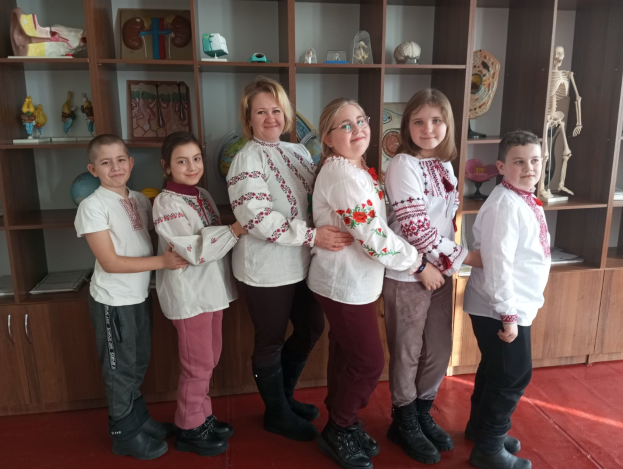 Gruppe von Kindern in traditioneller ukrainischer Kleidung vor einem Bücherregal mit Spielzeug, einem Skelett und anderen Gegenständen in einer ukrainischen Schulumgebung.