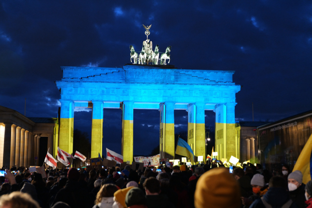 Eine Menge steht vor dem Brandenburger Tor in Berlin und hält Fahnen und Schilder, auf denen ein Banner mit protestrelevanten Text steht.