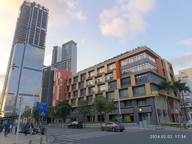 Eine belebte Stadtstraße mit hohen Gebäuden, Fahrzeugen, Passanten, Bäumen und einem Büroraggebäude mit grünem Dach unter einem bewölktem Himmel.