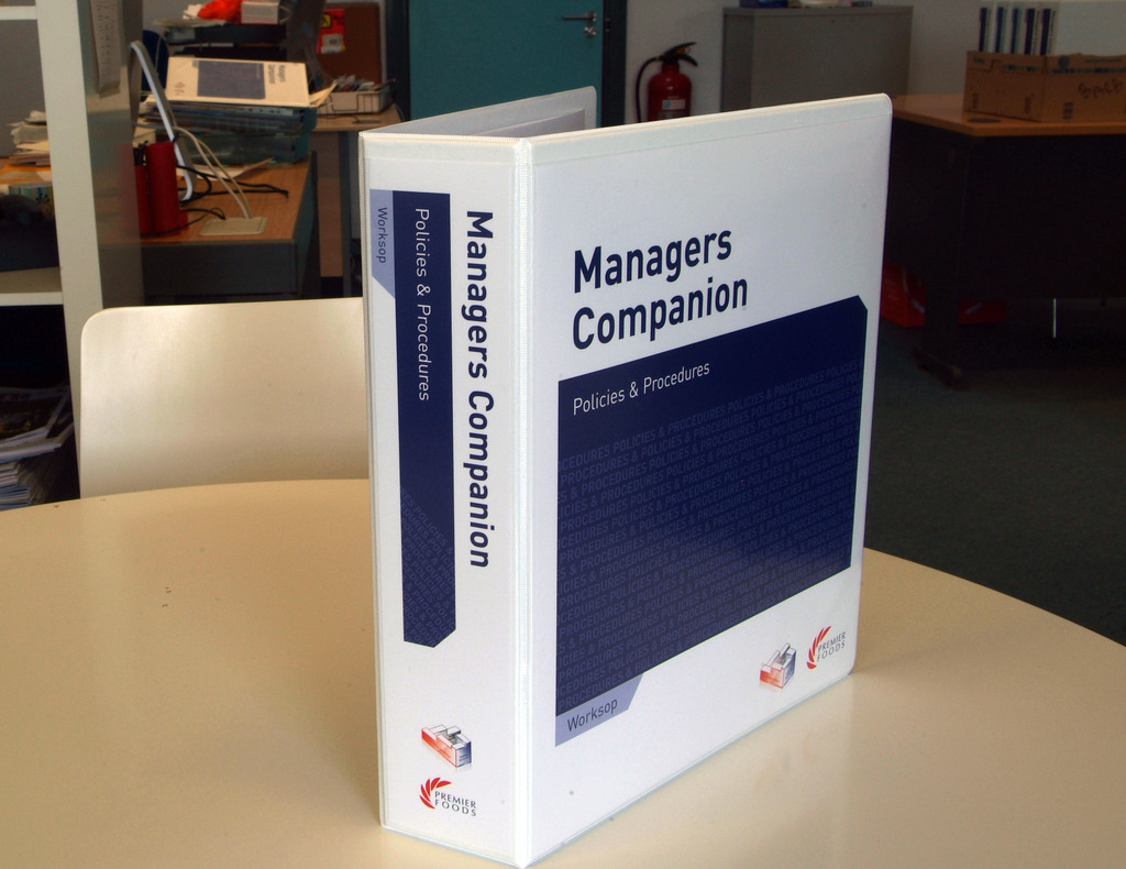 Ein Buch auf einem Tisch mit einem Stuhl dahinter, in der N├Ąhe einer T├╝r, Feuerl├Âscher, Akten und andere Gegenst├Ąnde.