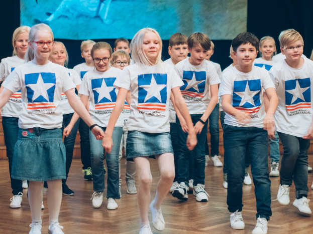 Gruppe von Kindern in weißen Hemden und blauen Shorts, die auf einem Holzboden vor einer Wand mit einem Bildschirm tanzen, an einem kunstnädler Tanzwettbewerb teilnehmen.