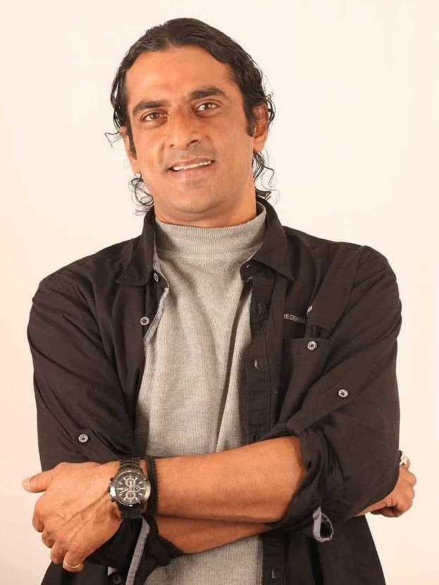 Ein Mann in einem schwarzen und aschfarbenen Kleid mit verschränkten Armen, der eine Uhr trägt und gegen einen weißen Hintergrund steht, sieht nachdenklich aus.