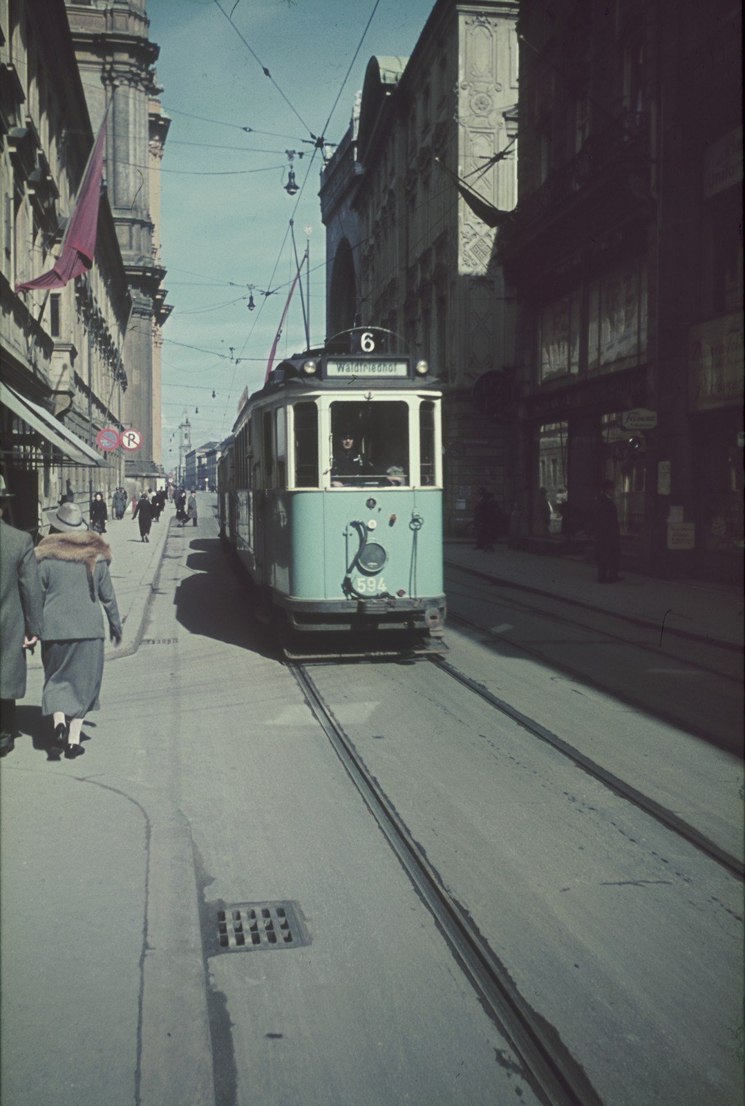 Altes Schwarz-Weiß-Foto einer Straßenbahn, die auf einer Stadtstraße mit Fußgängern auf dem linken Gehweg, Gebäuden und Strommasten im Hintergrund unter einem klaren Himmel fährt.