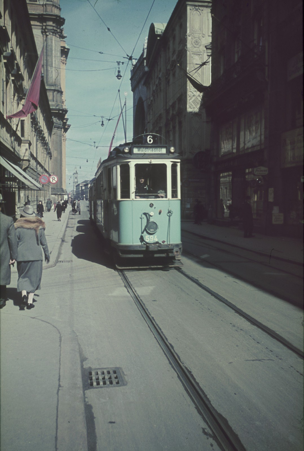 Altes Schwarz-Weiß-Foto einer Straßenbahn, die auf einer Stadtstraße mit Fußgängern auf dem linken Gehweg, Gebäuden und Strommasten im Hintergrund unter einem klaren Himmel fährt.
