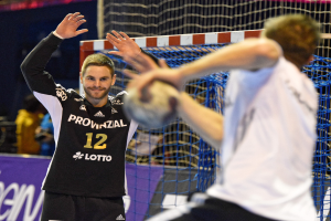Zwei Männer spielen Handball vor einem Tor mit Netz und Banner im Hintergrund und einigen Zuschauern in einer Arena.