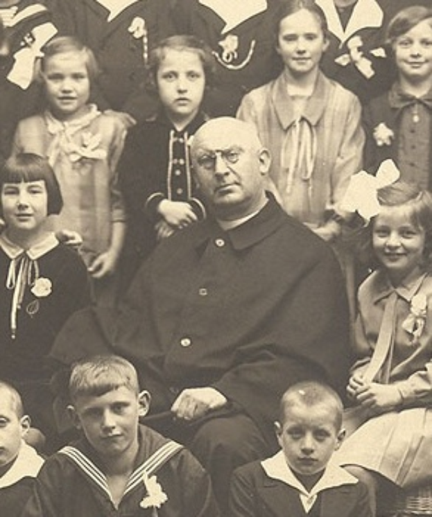 Ein altes Schwarz-Wei├č-Foto eines strengen Mannes, der in der Mitte sitzt und von einer Gruppe Kinder bewundert wird.