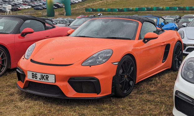 Ein Porsche 718 Boxster GTS auf der Goodwood Festival of Speed mit anderen Autos, einem Banner, Bäumen und einem klaren blauen Himmel im Hintergrund.
