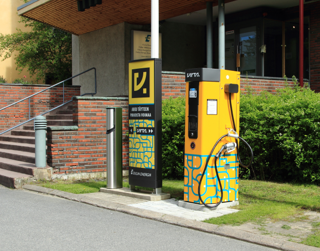 Eine Elektrofahrzeug-Ladestation am Straßenrand, umgeben von Gras, Pflanzen, Bäumen und einem Gebäude mit Fenstern und Säulen, mit Treppen mit Geländern, die zur Station führen.