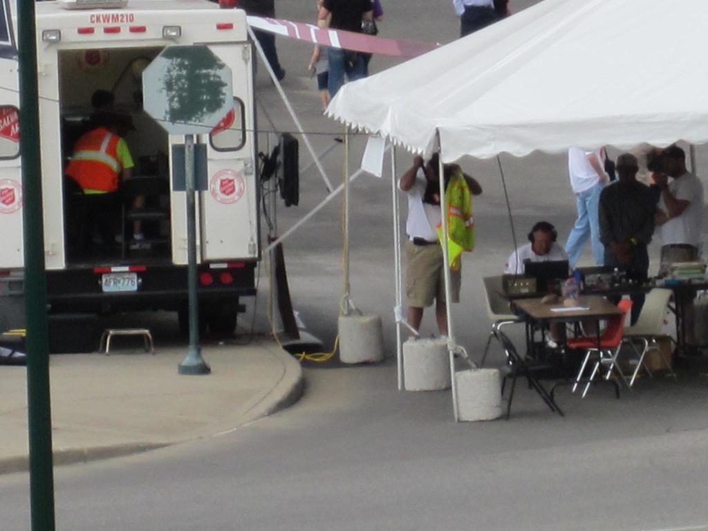 Ein Krankenwagen steht links von einer Stra√üe, w√§hrend auf der rechten Seite ein Zelt eine Gruppe von Menschen sch√ľtzt, mit einem Tisch und einem Stuhl in der N√§he.