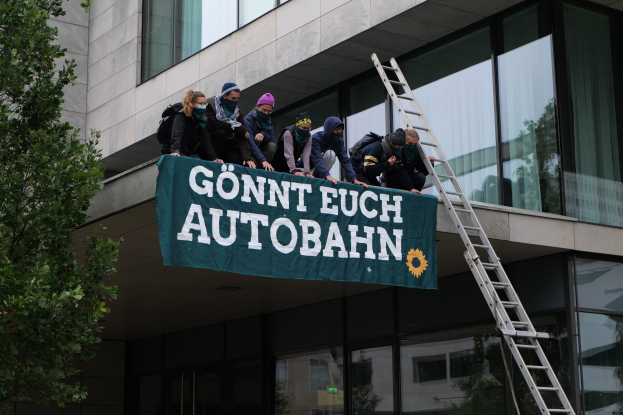 Eine Gruppe von Menschen auf einem Gebäude, das eine Fahne hält, mit einem Baum auf der linken Seite und Glasfenstern im Hintergrund, die scheinbar protestieren und die deutsche Regierung unterstützen.