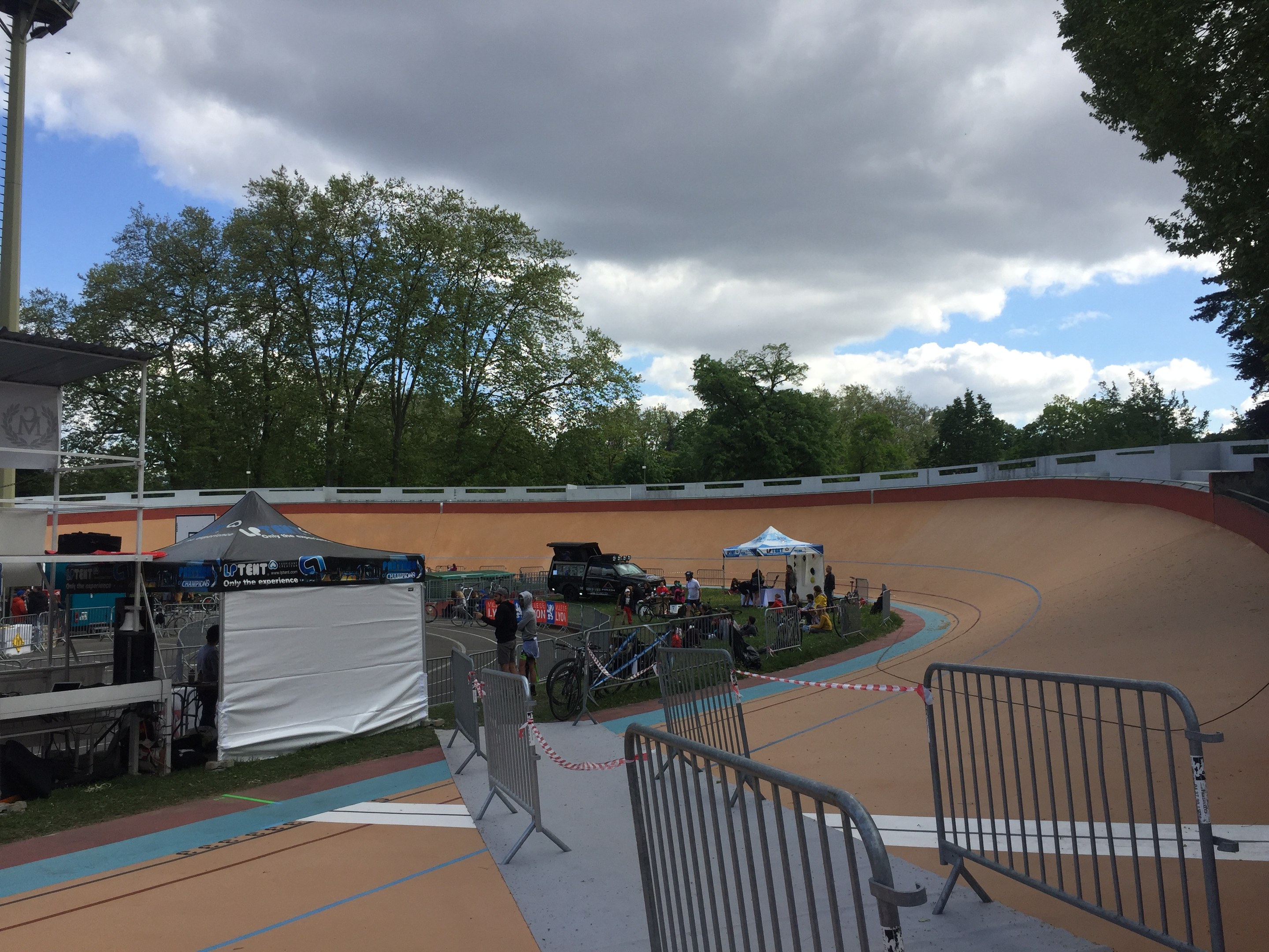 Gruppe von Radfahrern, die BMX-Räder auf einer Schmutzspur bei einem Outdoor-Event mit Zuschauern, Zelten und Fahrzeugen im Hintergrund fahren.