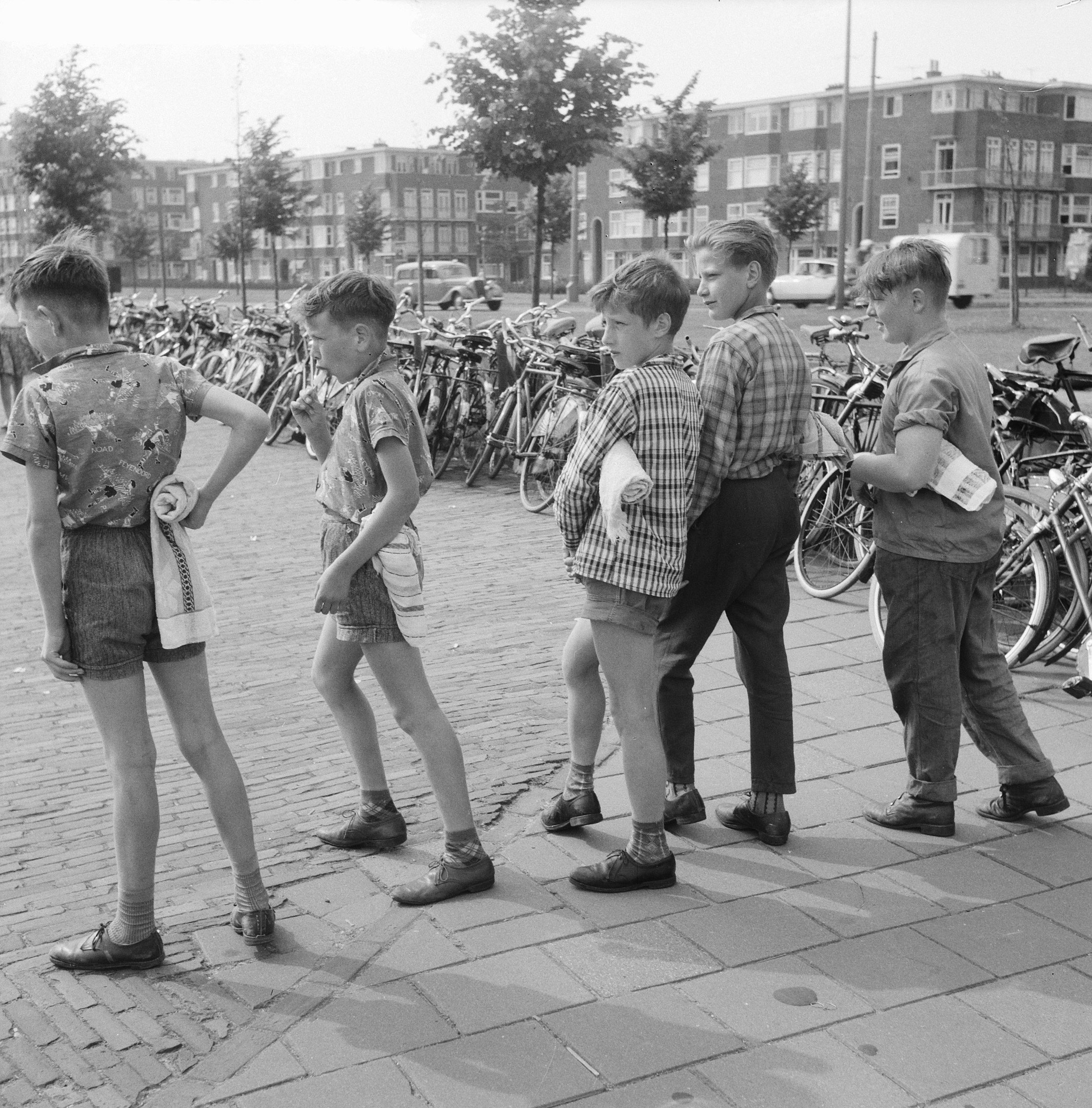 Eine Gruppe junger Jungs steht nebeneinander auf einem Bürgersteig, umgeben von Fahrrädern, Fahrzeugen, Bäumen, Pfählen und Gebäuden im Hintergrund, auf einem Schwarz-Weiß-Bild.