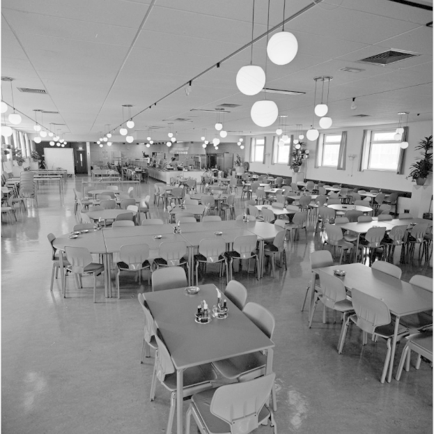 Große Kantine mit Tischen und Stühlen in Reihen, Flaschen und Gegenstände auf den Tischen, Deckenleuchten, Fenstern auf der rechten Seite und verstreuten Pflanzen.