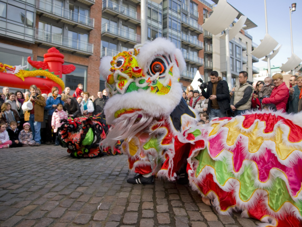 Ein farbenfrohes chinesisches Neujahrsfest in Amsterdam mit einem Drachen tanzen und einer Menge Schaulustiger, einige halten Kameras, vor einem Hintergrund aus Gebäuden, Laternenmästen und einem klaren blauen Himmel.