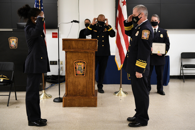 Eine Gruppe uniformierter Personen in Masken steht vor einem Podium mit einem Mikrofon, umgeben von Fahnen, Stühlen und Tischen, während einer Vereidigung eines neuen Feuerwehrchefs.