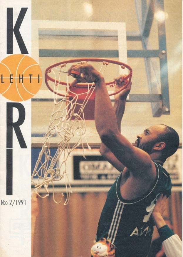 Ein Mann in einem Basketballtrikot wirft einen Ball durch den Korb mit einem Netz im Hintergrund, während eine weitere Person in der Nähe steht und eine Wand dahinter zu sehen ist.