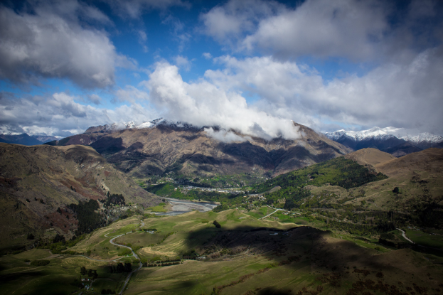 Panoramischer Blick von der Spitze eines Berges in Queenstown, Neuseeland, mit saftig grünem Gras, verstreuten Bäumen und einer gewundenen Straße unter einem Himmel voller weißer, flauschiger Wolken.