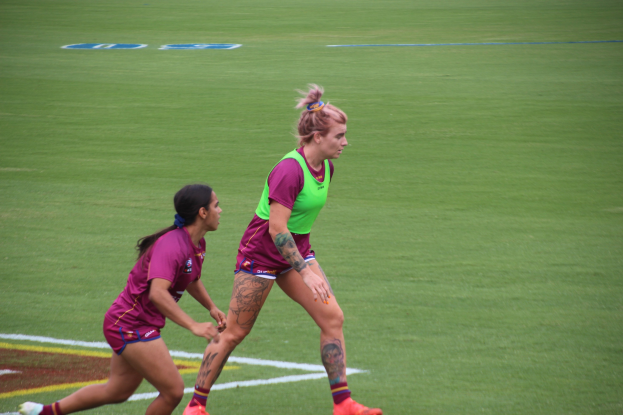 Zwei Frauen in Sportkleidung beim Fußballspielen auf einem Feld, eine in einem grünen T-Shirt und Shorts, die andere in einem pinken Kleid, beide mit sichtbaren Tattoos an Händen und Füßen.