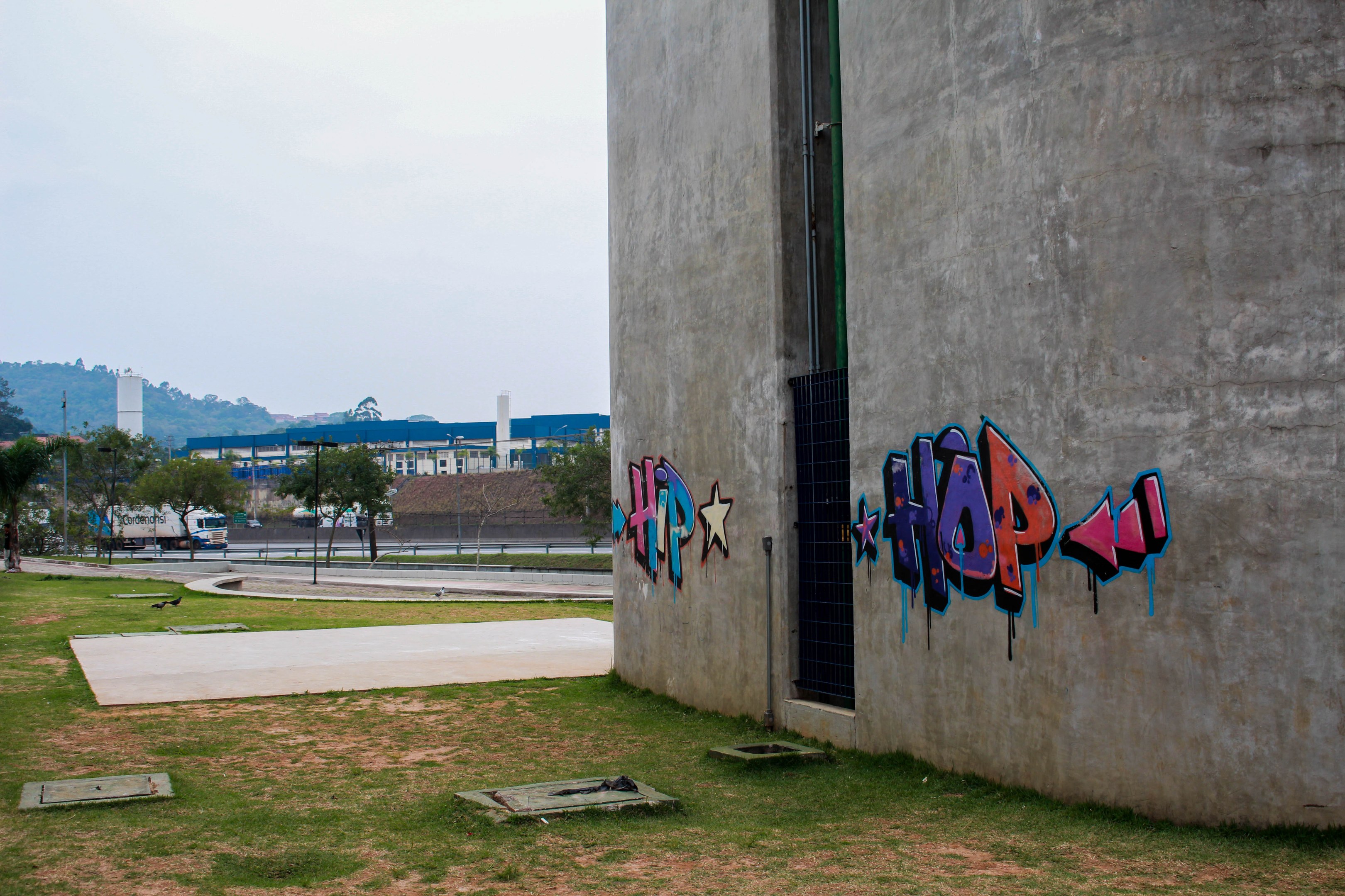 Betongebäude mit Graffiti, umgeben von Gras, Bäumen, Strommasten, Fahrzeugen auf der Straße, anderen Gebäuden, Hügeln und einem klaren blauen Himmel.