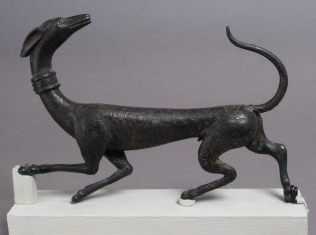 Eine Bronzestatuette eines Windhunds auf einem weißen Sockel vor einem grauen Hintergrund, mit detaillierter Darstellung des Fells und der Merkmale des Hundes.