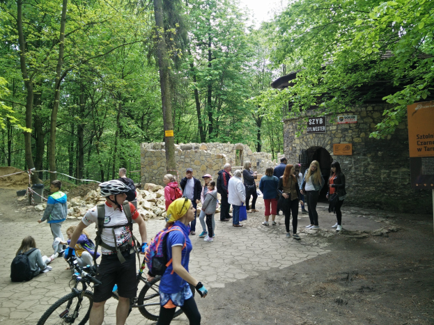 Gruppe von Menschen, die einen bewaldeten Pfad entlanggehen, einige tragen Helme und tragen Taschen oder Fahrräder, mit einer Steinmauer und einem Schild im Hintergrund, das den Eingang zum Szyb Stylweste Mountainbike-Trail anzeigt.