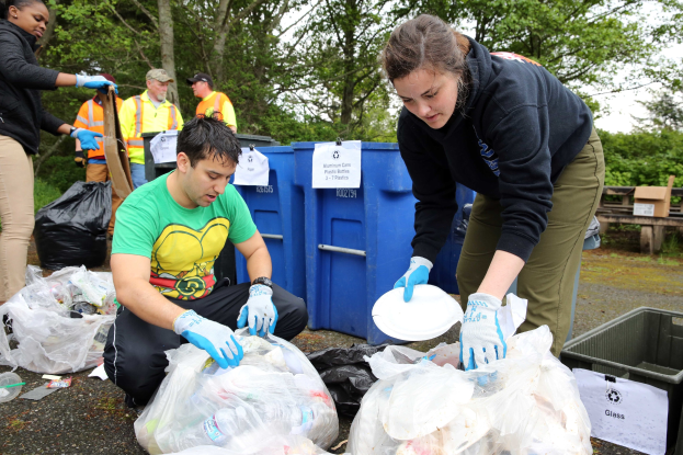 Eine Gruppe von Menschen, die Müll in einem Park sammeln, mit einem Mann und einer Frau in der Mitte, die Handschuhe tragen und Schilder halten, umgeben von Plastikabdeckungen, Flaschen und anderen Gegenständen auf dem Boden, einem Mülleimer und einer Holzbank auf der rechten Seite und Bäumen und einem klaren blauen Himmel im Hintergrund.