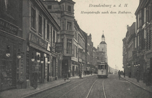 Schwarz-weiß-Foto einer Straßenkreuzung in Brandenburg, Deutschland mit einer Tram, Fußgängern, Gebäuden, Schildern, Straßenlaternen und einem bewölkten Himmel.