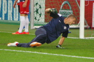 Ein Fussballspieler in Handschuhen taucht auf, um einen Ball vor einem Tor zu fangen, mit zwei Personen, einem Netz und einer Banner mit Text im Hintergrund.