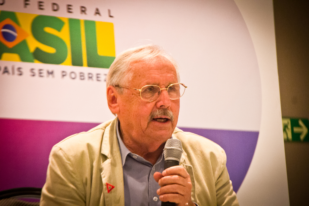 Eine Person, die in ein Mikrofon spricht und im Hintergrund ein Banner mit der Aufschrift 'Bundes' zu sehen ist.